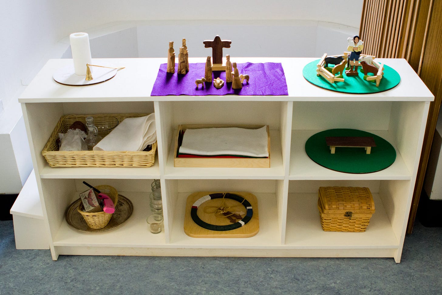 A Godly Play focal shelf with the Christ candle, Holy Family, and Good Shepherd on the top shelf, and baptism, circle of the church year, underlays, and other story pieces underneath.