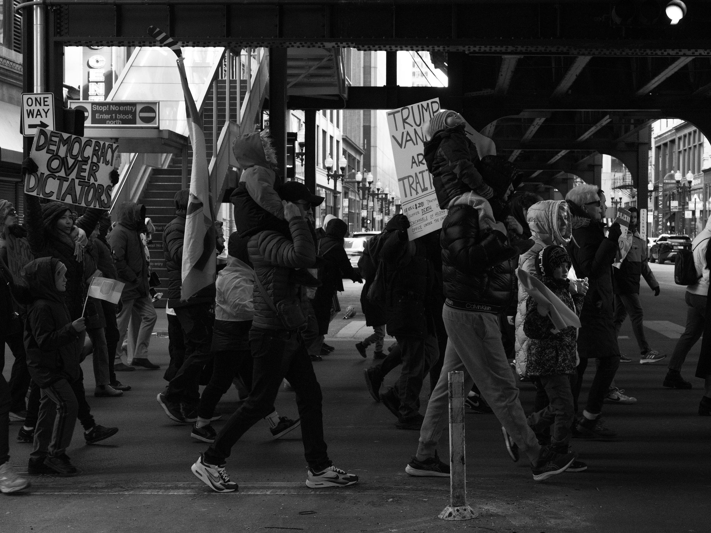 A black and white photograph captures a protest march moving beneath an elevated train track in a city. Demonstrators hold signs, including one that reads "Democracy Over Dictators" and another stating "Trump & Vance Are Traitors." Ukrainian flags wave among the crowd as parents carry children on their shoulders. The urban setting, with street signs, metal staircases, and traffic in the background, frames the marchers in their determined procession. A black and white photograph captures a protest march moving beneath an elevated train track in a city. Demonstrators hold signs, including one that reads "Democracy Over Dictators" and another stating "Trump & Vance Are Traitors." Ukrainian flags wave among the crowd as parents carry children on their shoulders. The urban setting, with street signs, metal staircases, and traffic in the background, frames the marchers in their determined procession.