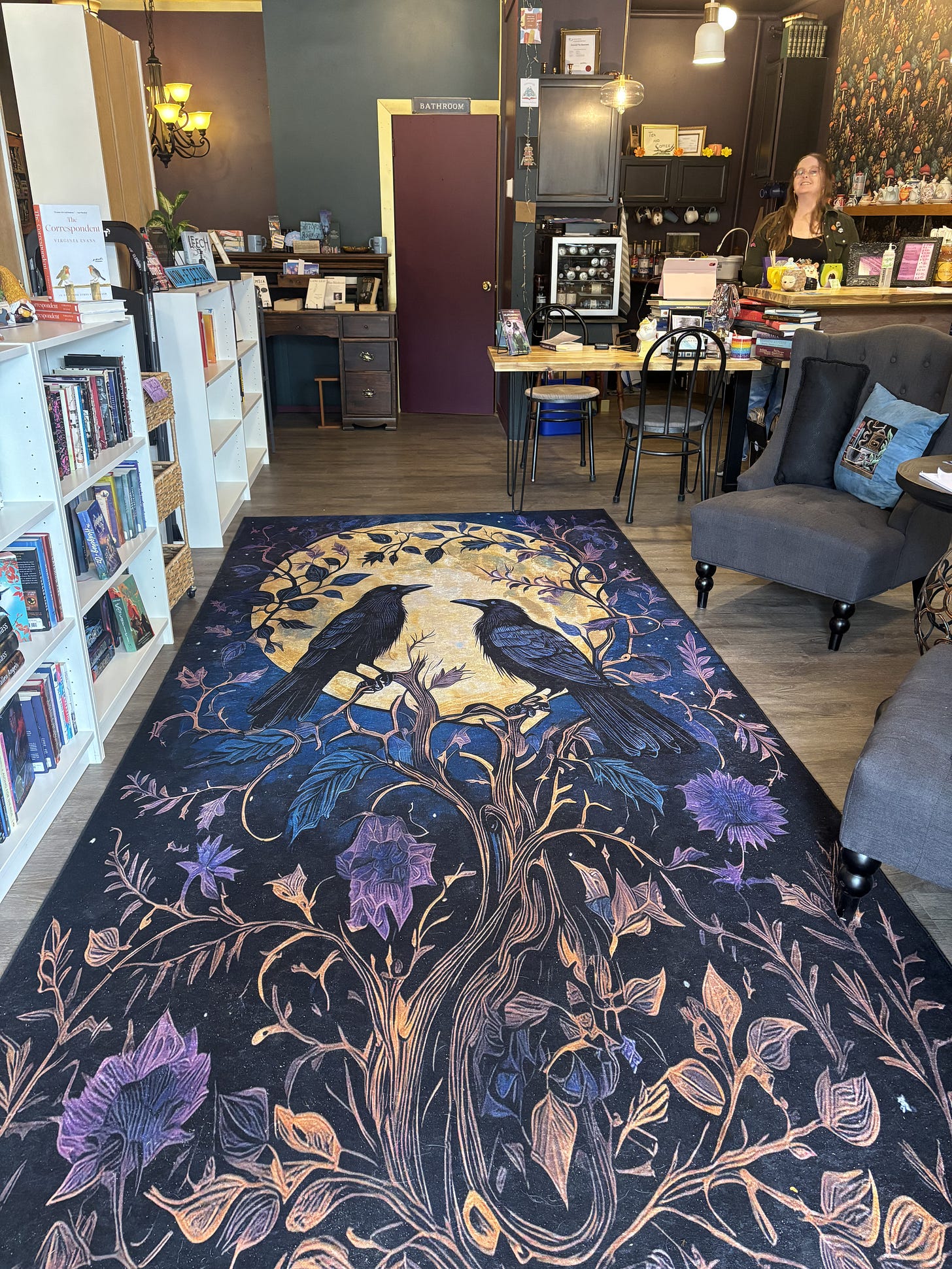 The entry to a bookstore features a large rug with two ravens in black sitting on brown tree branches in front of a yellow moon. A woman stands in the back of the photo, and books line the left side. Two armchairs with pillows are on the right.