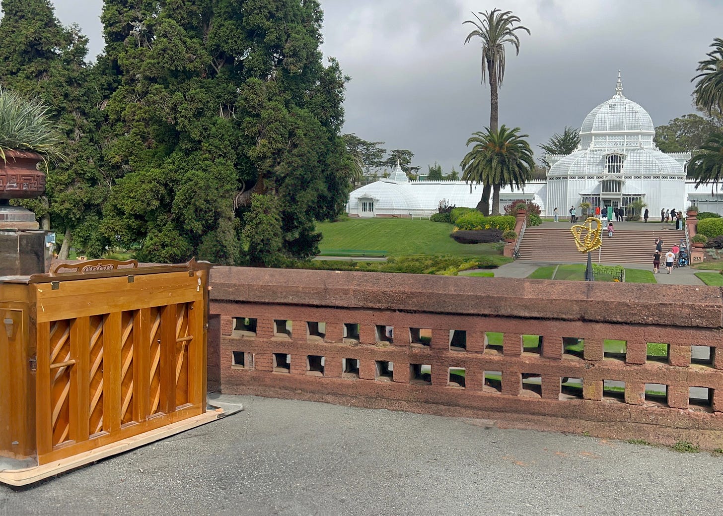 A photo at San Francisco's Conservatory of Flowers under overcast skies with tall palms and cypress trees framing the iconic white glass-domed greenhouse in the background. In the foreground on a concrete path near a red brick wall with geometric cutouts stands a polished wooden grand piano on its side, positioned beside a large terracotta planter with agave plants. A photo at San Francisco's Conservatory of Flowers under overcast skies with tall palms and cypress trees framing the iconic white glass-domed greenhouse in the background. In the foreground on a concrete path near a red brick wall with geometric cutouts stands a polished wooden grand piano on its side, positioned beside a large terracotta planter with agave plants.