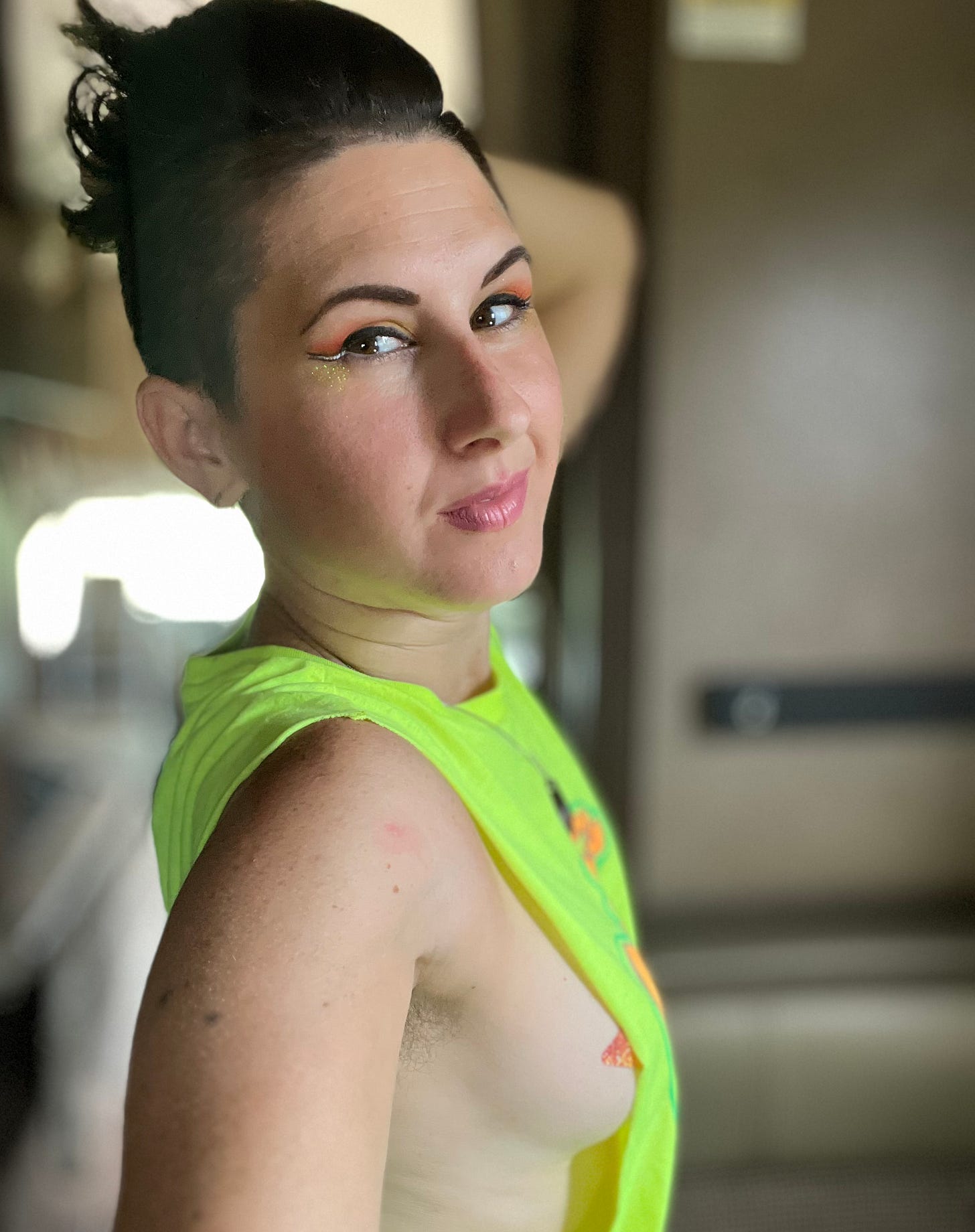 Lyric Rivera, in a neon yellow shirt that shows a lot of their bare side, but a pastie is covering the “vital parts” - they have gold and orange winged eyeliner, pink lipstick, and are posing for a selfie inside their RV. Lyric Rivera, in a neon yellow shirt that shows a lot of their bare side, but a pastie is covering the “vital parts” - they have gold and orange winged eyeliner, pink lipstick, and are posing for a selfie inside their RV.
