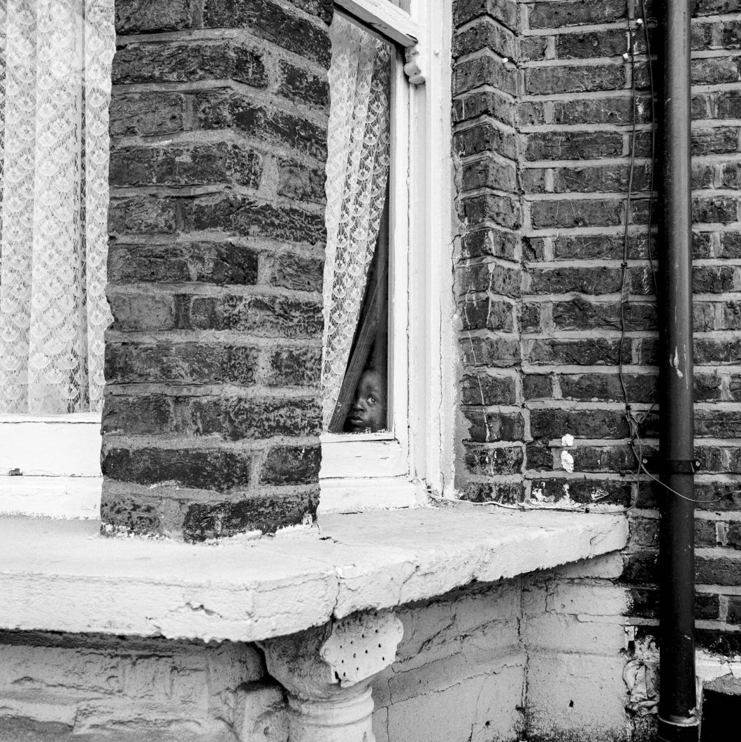 Black and white photo of a typical early twentieth century terrace house bay window with white net curtains in the window. In the side of one pane, a small Black boy has peeled the curtain to side and peeps out. The photographer has captured the moment beautifully.