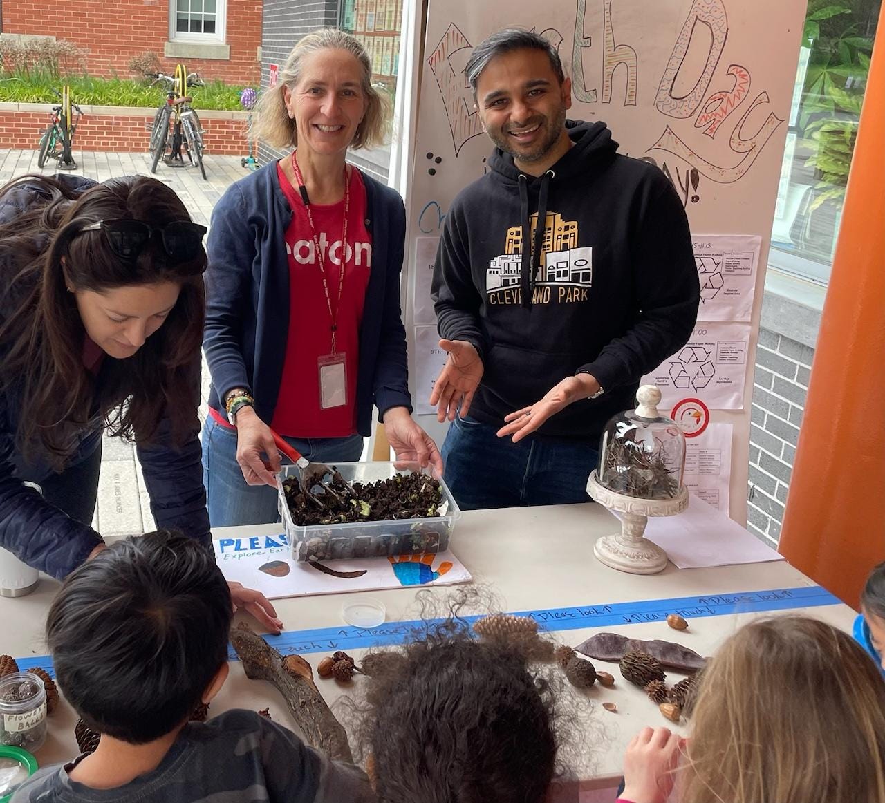 Picture of Sauleh gesturing to a box of compost that Eaton elementary school students are exploring. You can see the back of three heads of students. Sauleh is wearing a black sweatshirt that says Cleveland Park and is standing next to someone in a red Eaton shirt. They are standing at a table outside and a whiteboard sign behind Sauleh says "Earth Day" in handwriting. 