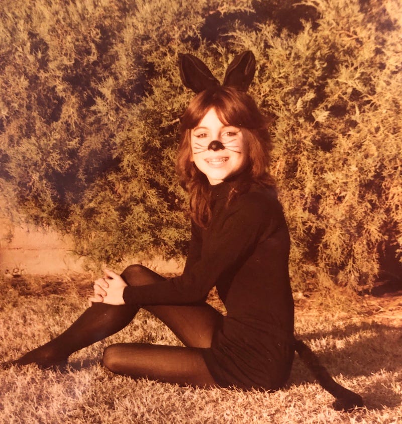A girl seated on dry grass, in all black, with cat ears, tail and painted whiskers