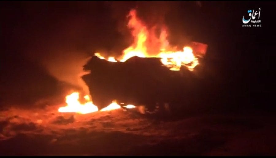 First image shows a nighttime scene with intense orange flames and thick black smoke rising from a structure or vehicle behind a wall, surrounded by trees and a lit road, with Arabic text overlay from Amaq News Agency. Second image depicts a close-up of a large vehicle engulfed in bright yellow and orange fire with heavy smoke billowing upward, Arabic watermark visible at the bottom. Both images capture the aftermath of an attack in a rural area with dark surroundings.