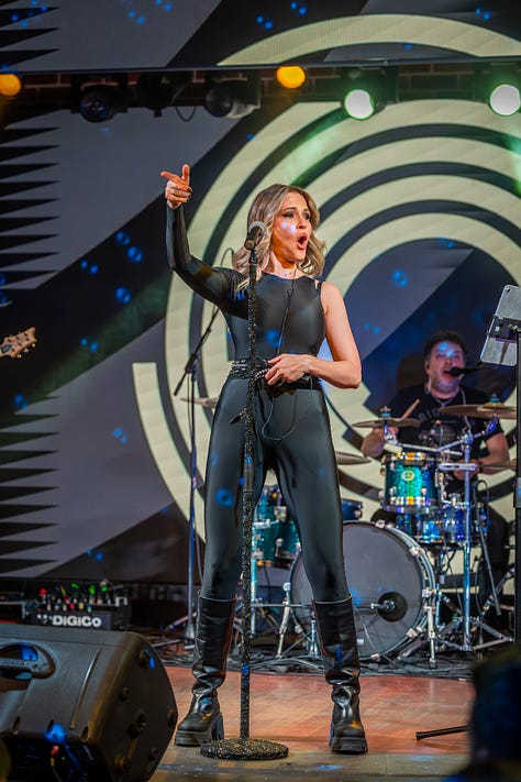 Alina Crișan & Keo doi artiști cântă împreună pe scenă, față în față, fiecare ținând câte un microfon. Artista poartă o ținută neagră, iar artistul poartă o geacă din piele. Fundalul scenei are elemente grafice luminoase în tonuri de verde și galben, iar atmosfera este specifică unui concert live.