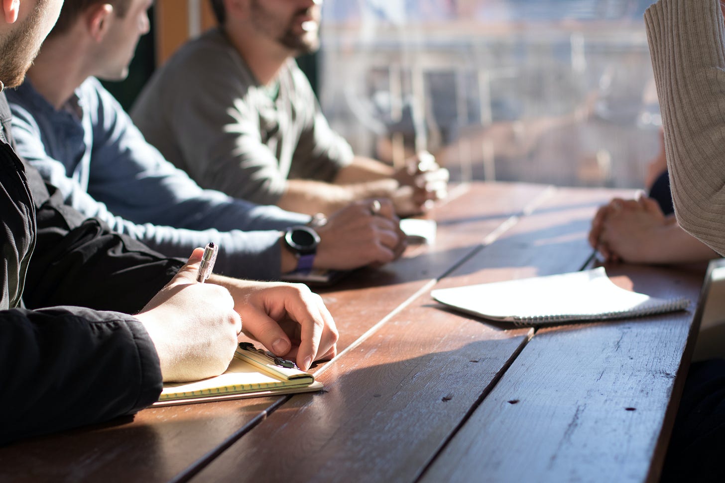 A photo of a group seated at a table taking notes on paper, listening to a speaker, also seated. A photo of a group seated at a table taking notes on paper, listening to a speaker, also seated.