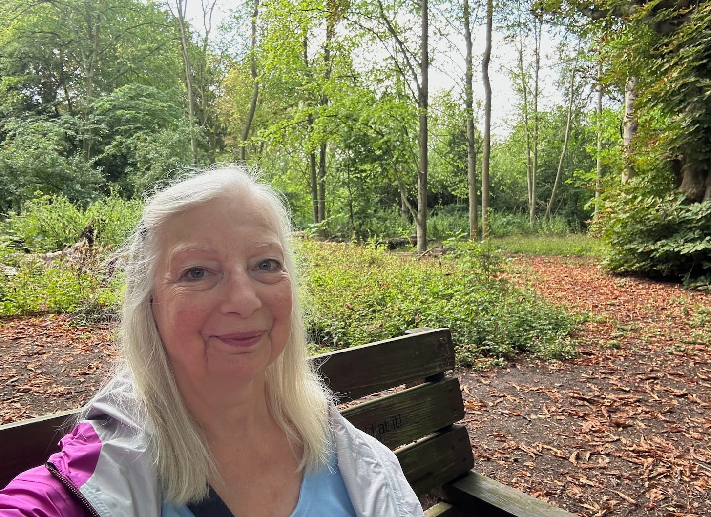 Woman with long grey hair sits on a wooden bench with trees in the background