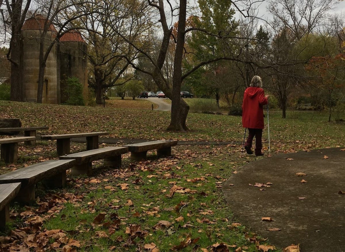 Woman walking with ski poles on an autumn day. She has grey hair and wears a red raincoat.