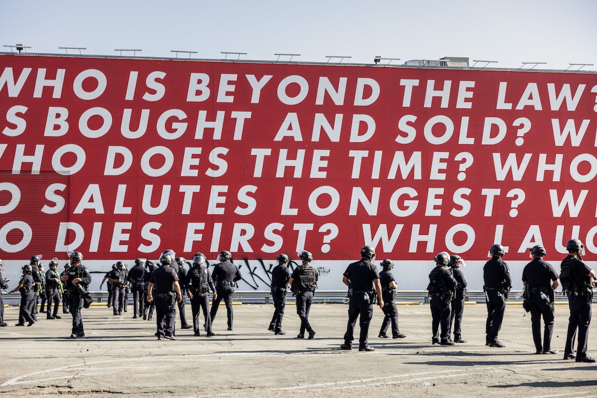 A group of soldiers beneath a red mural with text that begins 'WHO IS BEYOND THE LAW?' A group of soldiers beneath a red mural with text that begins 'WHO IS BEYOND THE LAW?'