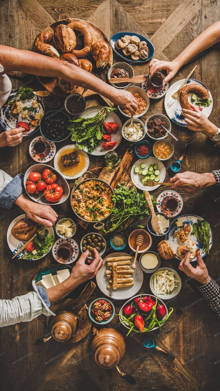 This may contain: a group of people sitting around a table filled with food and drinks, all holding their hands together