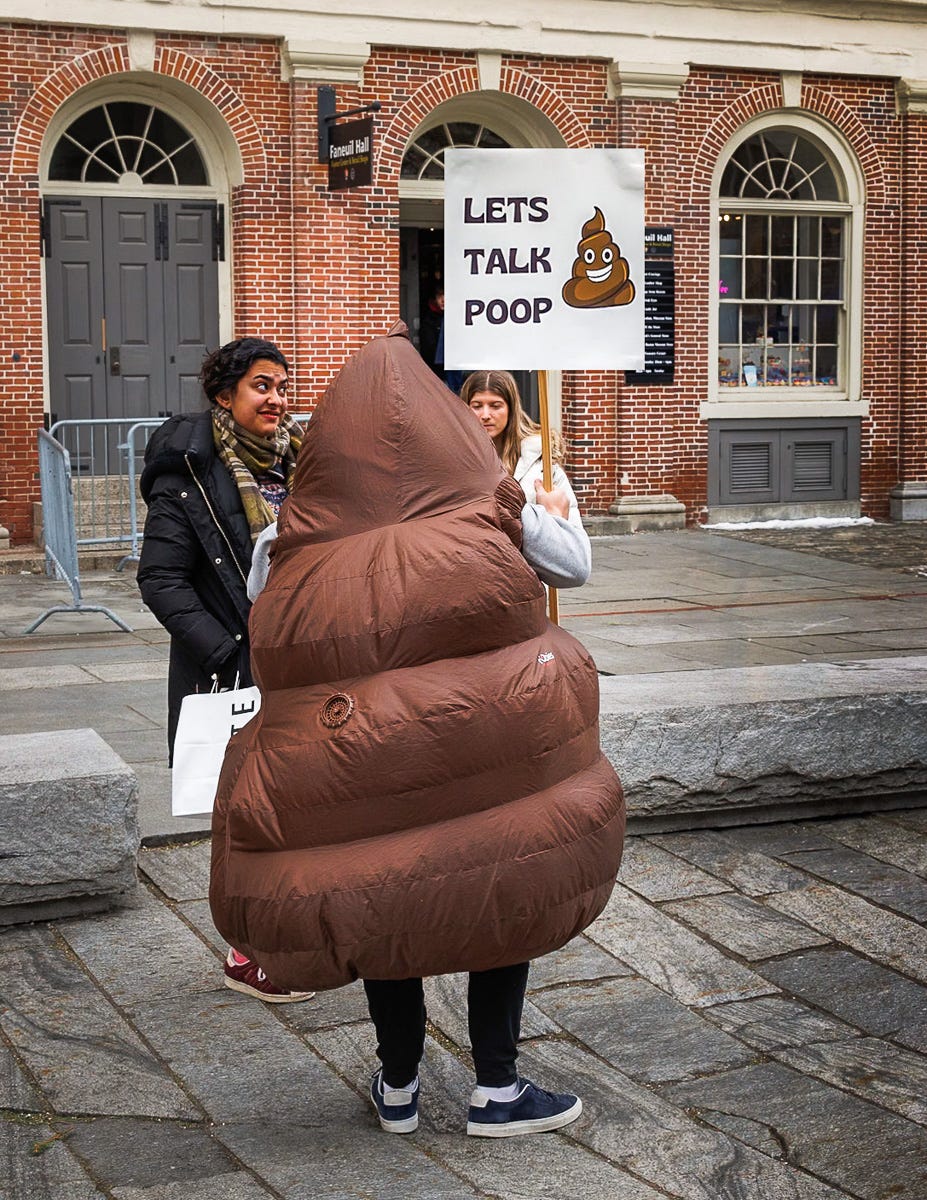 Person in a poop emoji costume holding a “Let’s Talk Poop” sign, engaging with two women in front of Faneuil Hall in Boston. Person in a poop emoji costume holding a “Let’s Talk Poop” sign, engaging with two women in front of Faneuil Hall in Boston.