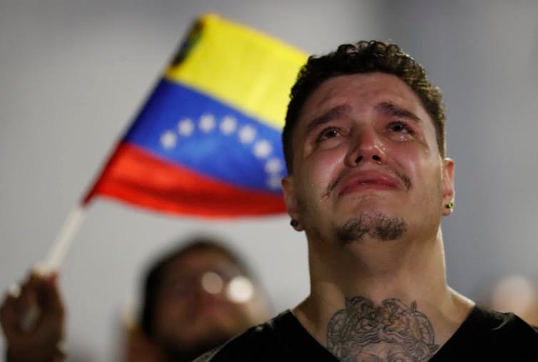 Un venezolano tatuado llora con alguien ondeando una bandera de Venezuela detrás de él.