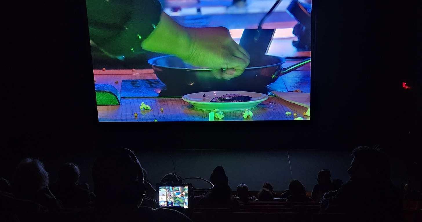 A photo of a dark theater, from a back row. On screen, a Black hand is doing something mysterious on a table, which has a plate and metal bowl and various detritus on it. In the foreground, a small screen is being held by an audience member; the screen is lit up, and one of the squares on the screen is the picture being projected on the theater screen.