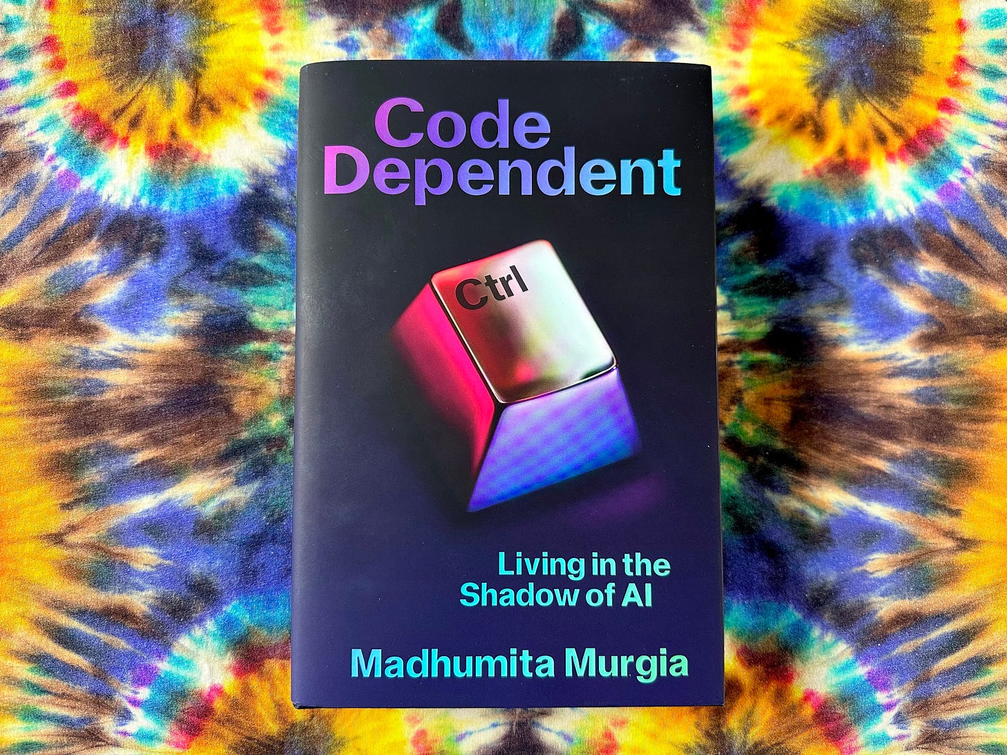 Picture of a book titled "Code Dependent", by Madhumita Murgia, on a multi-coloured tie-dye print cloth. The cover is dark navy with a giant close-up of a CTRL button with the title “Code Dependent: Living in the Shadow of AI”. Picture of a book titled "Code Dependent", by Madhumita Murgia, on a multi-coloured tie-dye print cloth. The cover is dark navy with a giant close-up of a CTRL button with the title “Code Dependent: Living in the Shadow of AI”.