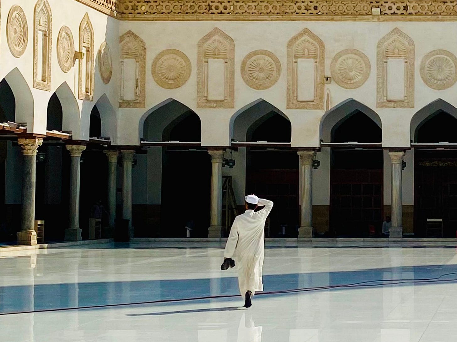 A man in a white robe and cap walks across the polished marble courtyard of Al-Azhar Mosque in Cairo, shielding his eyes, with carved arches and columns behind him.