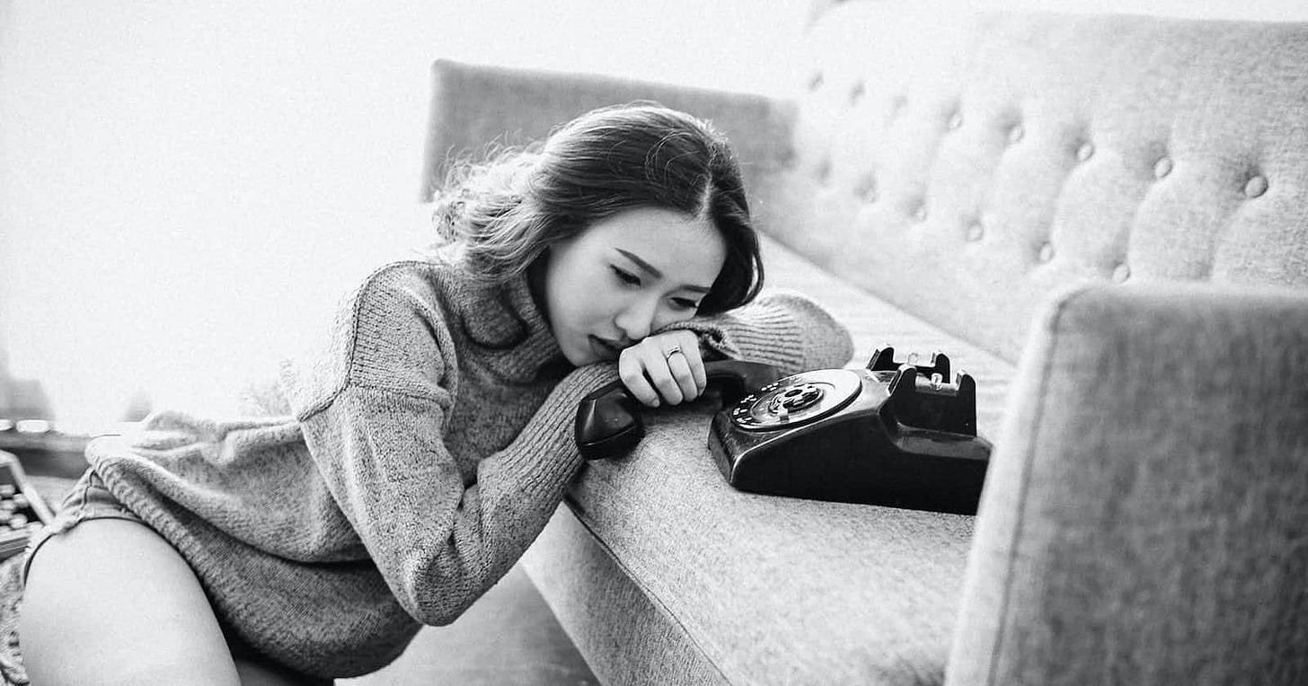 A sad woman sits on the floor by a couch, holding an old phone receiver, appearing dejected after an emotional call.