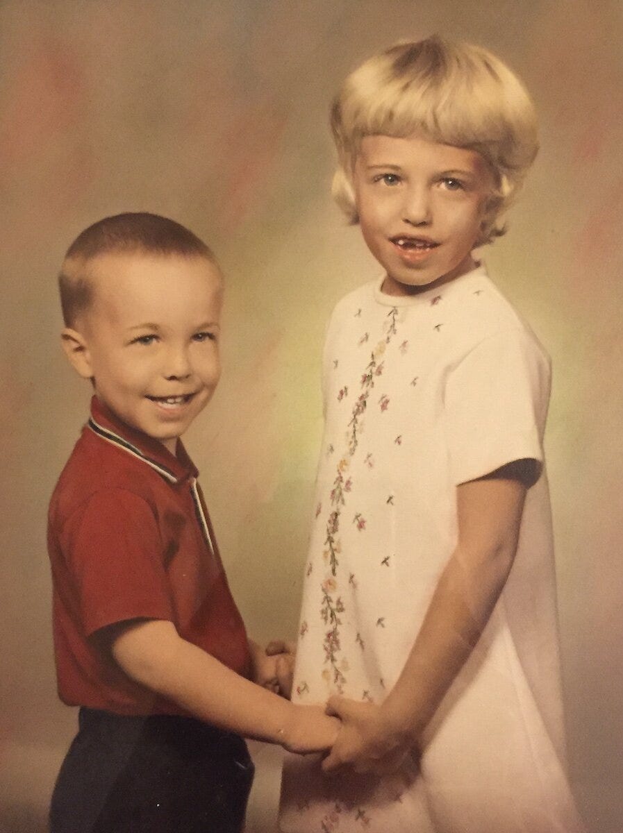 Studio photograph of boy about 6 and girl about 8 facing each other holding hands smiling at the camera