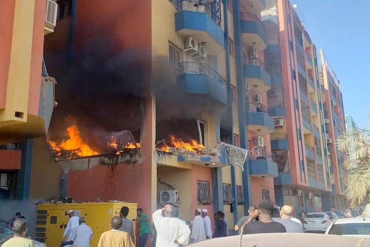 An apartment block bombed with fire burning and people watching from the street.
