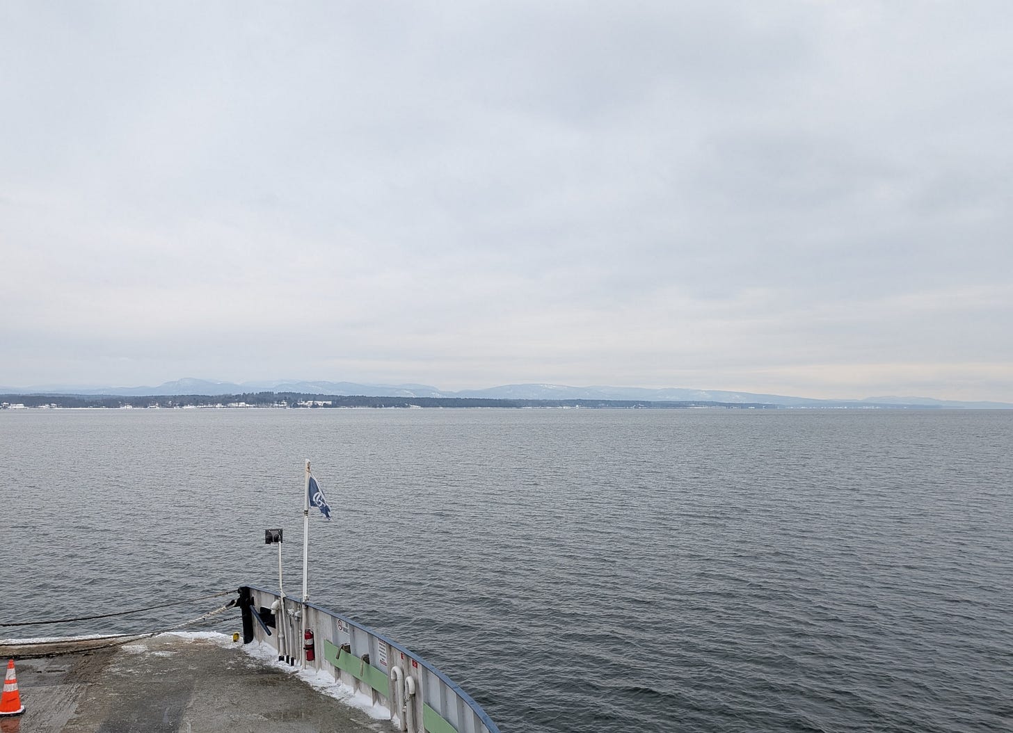 a view from a ferry boat. you can see the deck of the ferry with a blue flag waving in the wind. beyond is an expanse of water and mountains in the distance on the shore