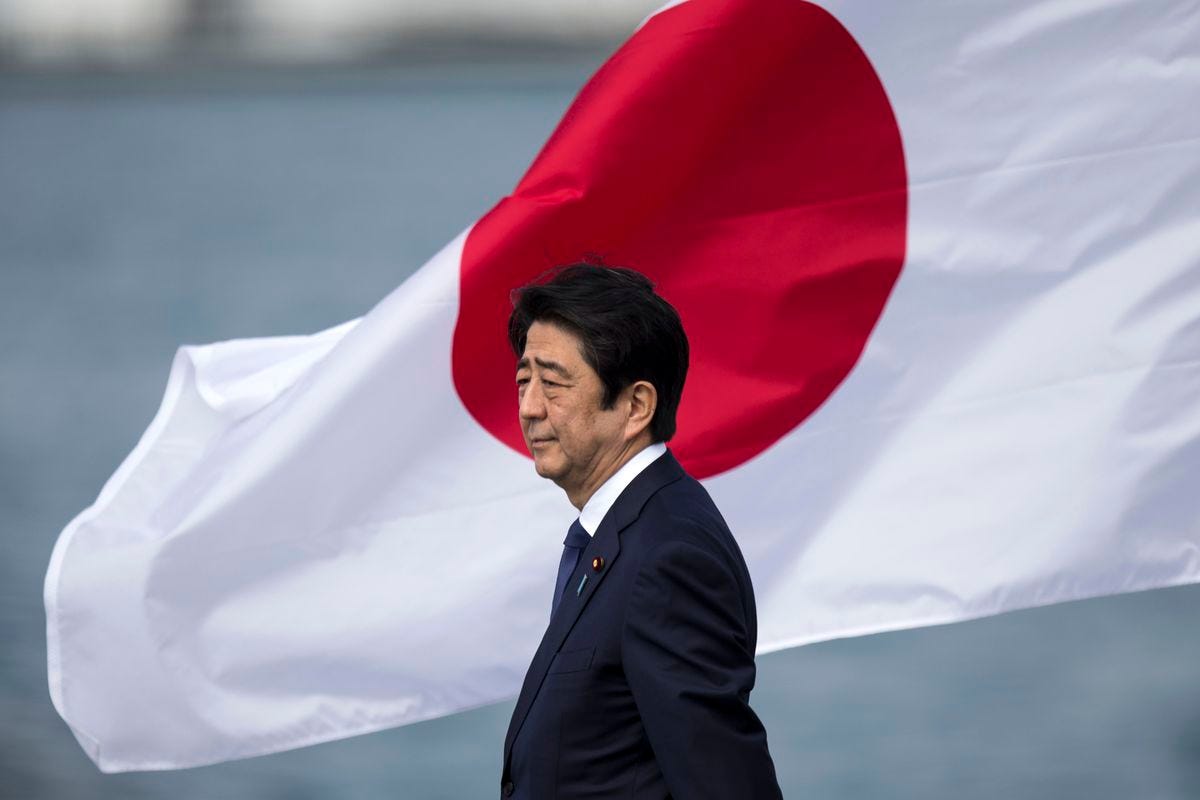 Shinzo Abe stands in a dark suit, in profile, against a massive Japanese flag furrowed by wind behind him. Shinzo Abe stands in a dark suit, in profile, against a massive Japanese flag furrowed by wind behind him.