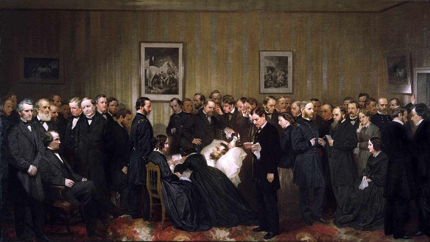 A crowded bedroom lit by lamps, with dozens of men in dark coats standing around a bed where Abraham Lincoln lies under a white sheet, a woman bent over him in grief.