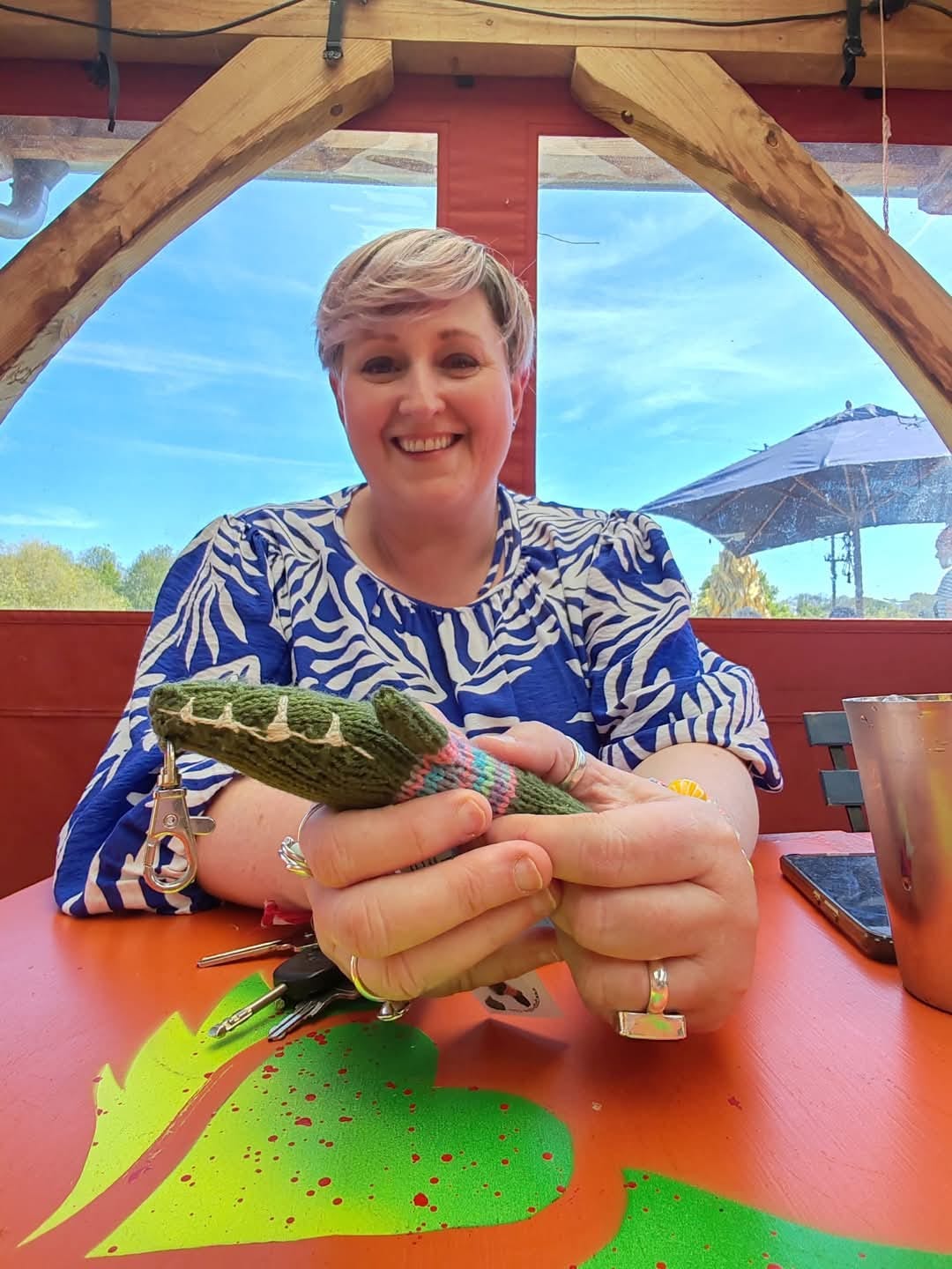 Jill Holly—shoulders and above—sat leaning on a table. She is white-skinned, short pale hair, smiling. Blue/white print dress. She’s holding some keys in her hands. Her hands are much closer to the camera lens and are out of proportion—much larger in scale, very comically so. Blue sky behind in the distance.