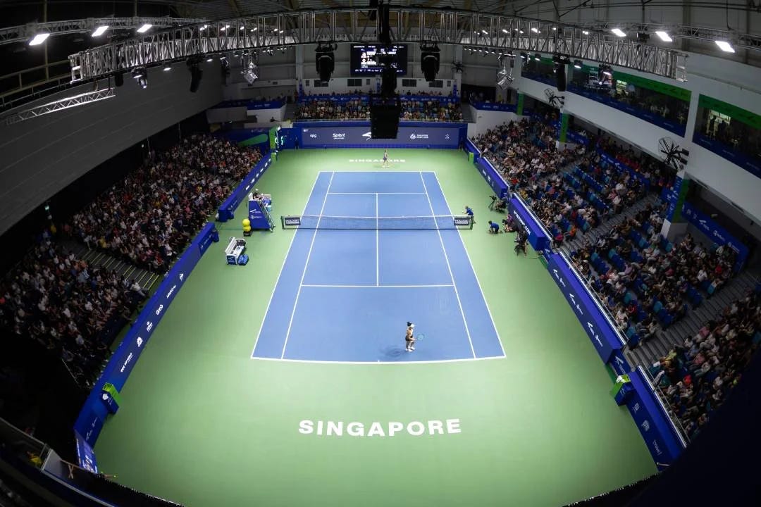 A high-angle, wide wide-shot photograph titled "SIngapore Tennis Open 2026 The Kallang.webp" capturing an indoor professional tennis match at the OCBC Arena. Two female players face each other across a vibrant blue hard court surrounded by green outer borders, with the word "SINGAPORE" printed in large white letters at the base of the court. The arena stands are filled with spectators under bright overhead stadium lighting rigs, visually capturing the atmosphere of the Singapore Tennis Open following its upgrade to a WTA 500 tournament.