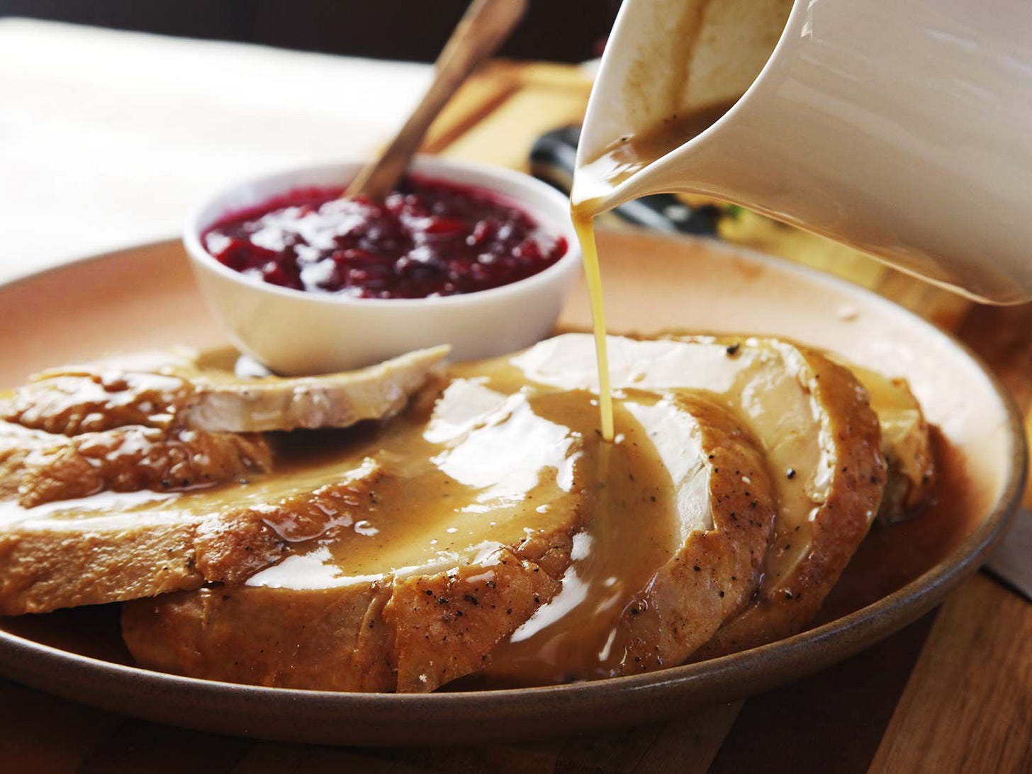 Pouring gravy from a pitcher over a platter of sliced roasted turkey breast meat, with a bowl of cranberry sauce in the background.