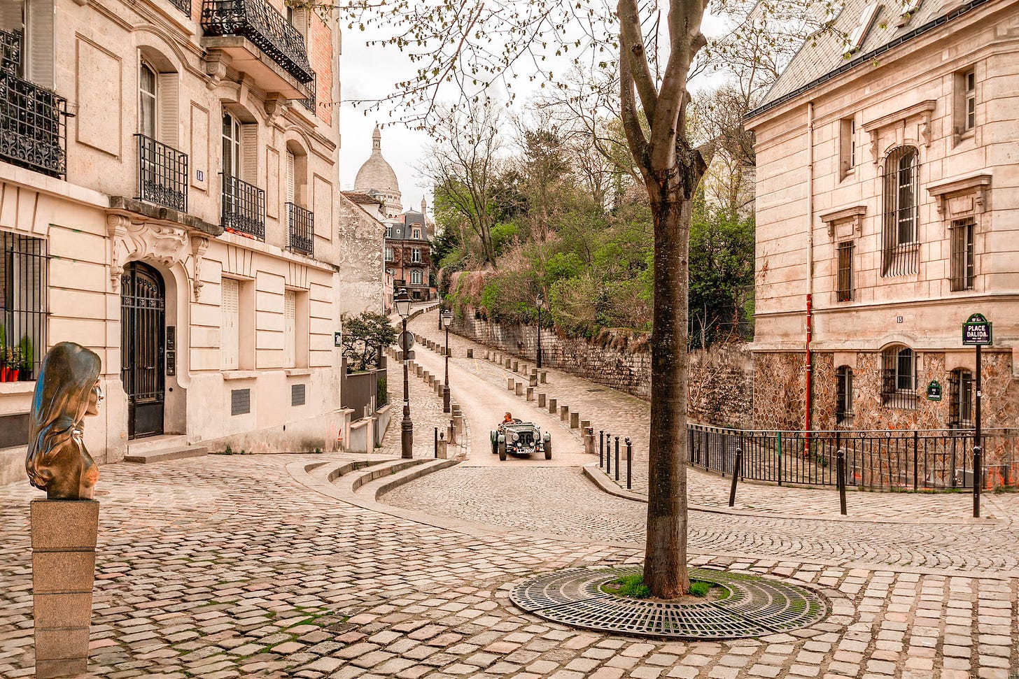 View from Place Dalida looking down Rue de l'Abreuvoir in Montmartre, with pastel houses and the domes of Sacré-Cœur rising above the rooftops in Paris.
