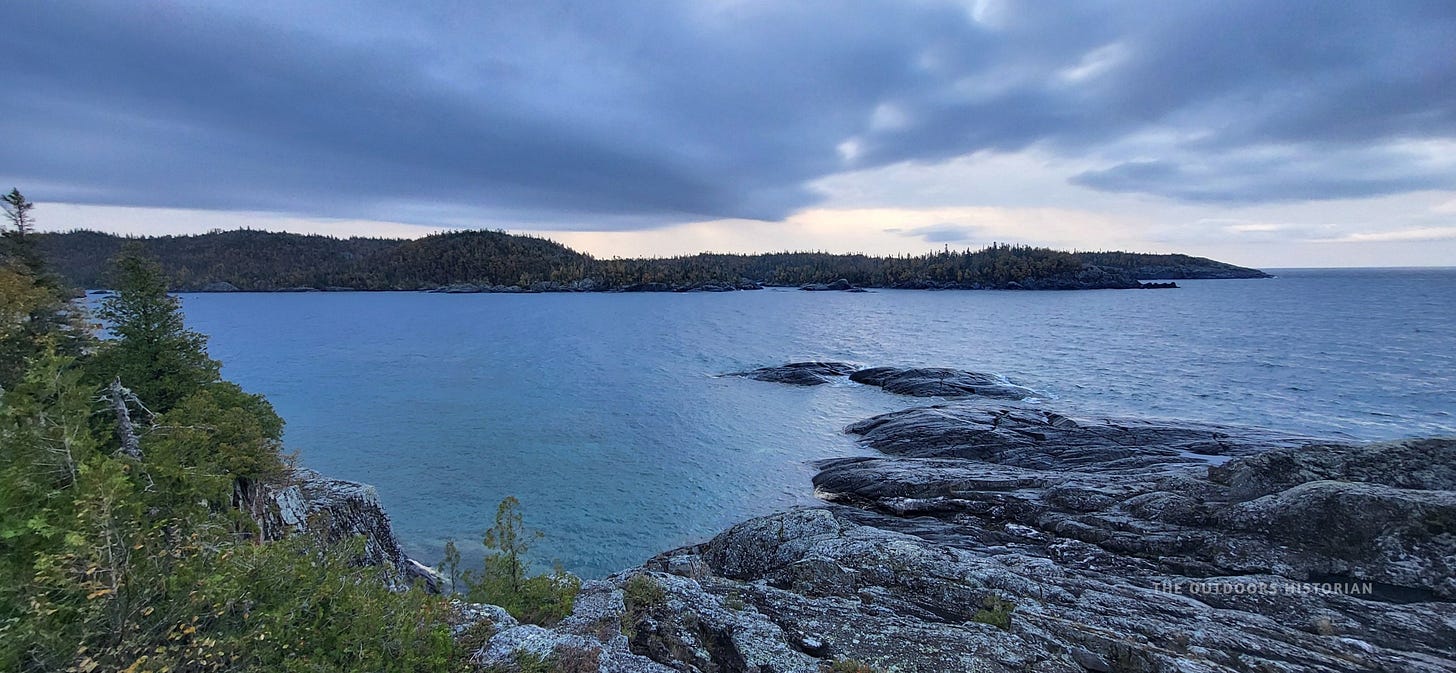 A rugged, rocky shoreline made of dark, textured granite. The sky above is moody and grey, with dramatic storm clouds rolling over the blue grey water of Lake Superior.