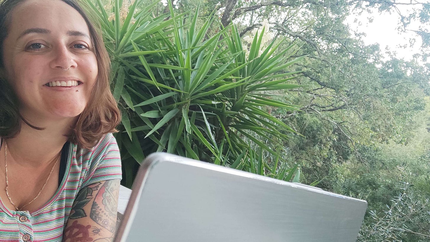 Sophie smiling at the camera while working on her silver laptop wth lots of lush green foliage behind her. Sophie smiling at the camera while working on her silver laptop wth lots of lush green foliage behind her.