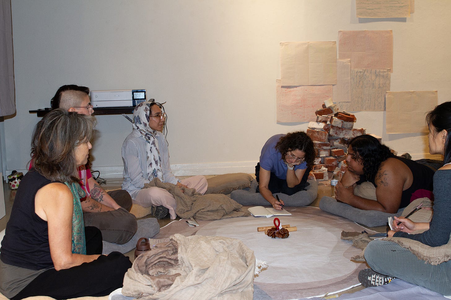 Seven people sit on cushions in a rough circle around a pale round rug. One person lies down at the edge of the circle. A small arrangement of objects sits at the rug's center. A stack of bricks rises behind them.