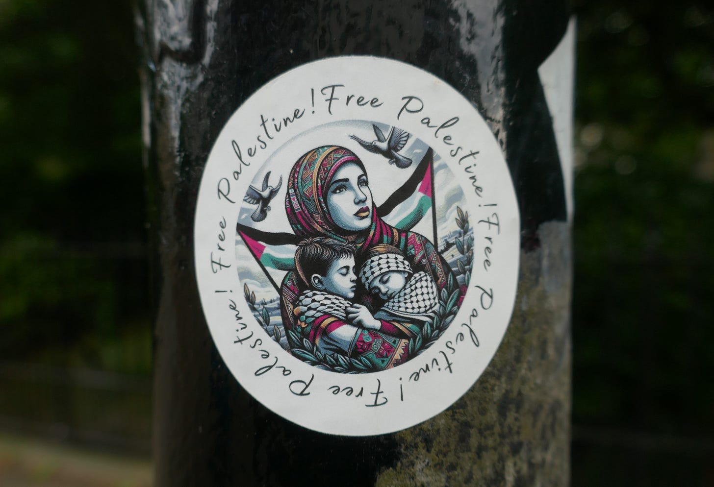 Photo of a sticker stuck on a tree that says "Free Palestine" and has artwork of a Palestinian person wearing a hijab and carrying two children wrapped in keffiyahs in their arms, with the Palestinian flag behind them and doves flying above
