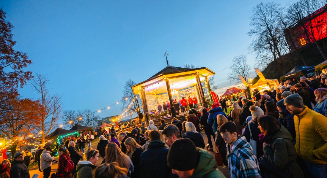 A crowd of people walking around a Christmas market, with a gazebo and a castle behind them and a blue sky above them