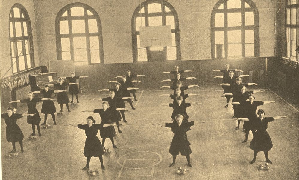 Rows of young women in matching uniforms stretch their arms out wide in rows in a school gymnasium. C. 1918 Rows of young women in matching uniforms stretch their arms out wide in rows in a school gymnasium. C. 1918