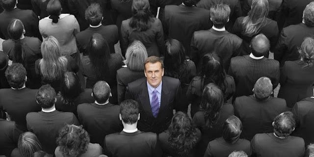 #descrevemospravocê  A imagem mostra um homem de meia-idade em pé, no centro de uma multidão de pessoas que estão todas vestidas de terno preto. A fotografia está em preto e branco, exceto pelo homem central, que aparece em cores. Ele tem cabelo castanho claro, penteado para o lado, pele clara e expressão séria, olhando diretamente para a câmera. Usa terno preto, camisa azul clara e uma gravata roxa com detalhes discretos, destacando-se visualmente do restante das pessoas ao redor. Os demais, vistos de costas ou de perfil, estão alinhados próximos uns dos outros, formando um padrão visual denso, todos usando ternos escuros, sem detalhes visíveis em seus rostos ou expressões, com cabelos em tons de cinza devido ao efeito da imagem em preto e branco. O homem colorido, em contraste, simboliza destaque e individualidade em meio a um grupo uniforme, em uma atmosfera corporativa, sugerindo o tema de se destacar na multidão ou liderança em ambientes profissionais.