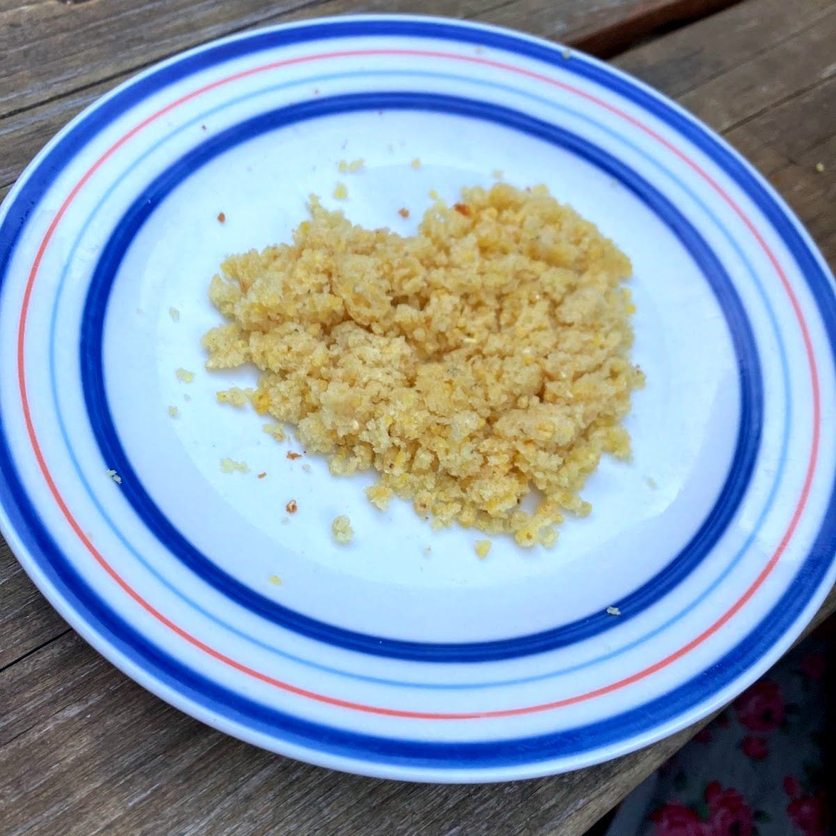 Photo of a heart shape made from golden cornbread crumbles on a white plate with blue and red striped borders, placed on a wooden surface.
