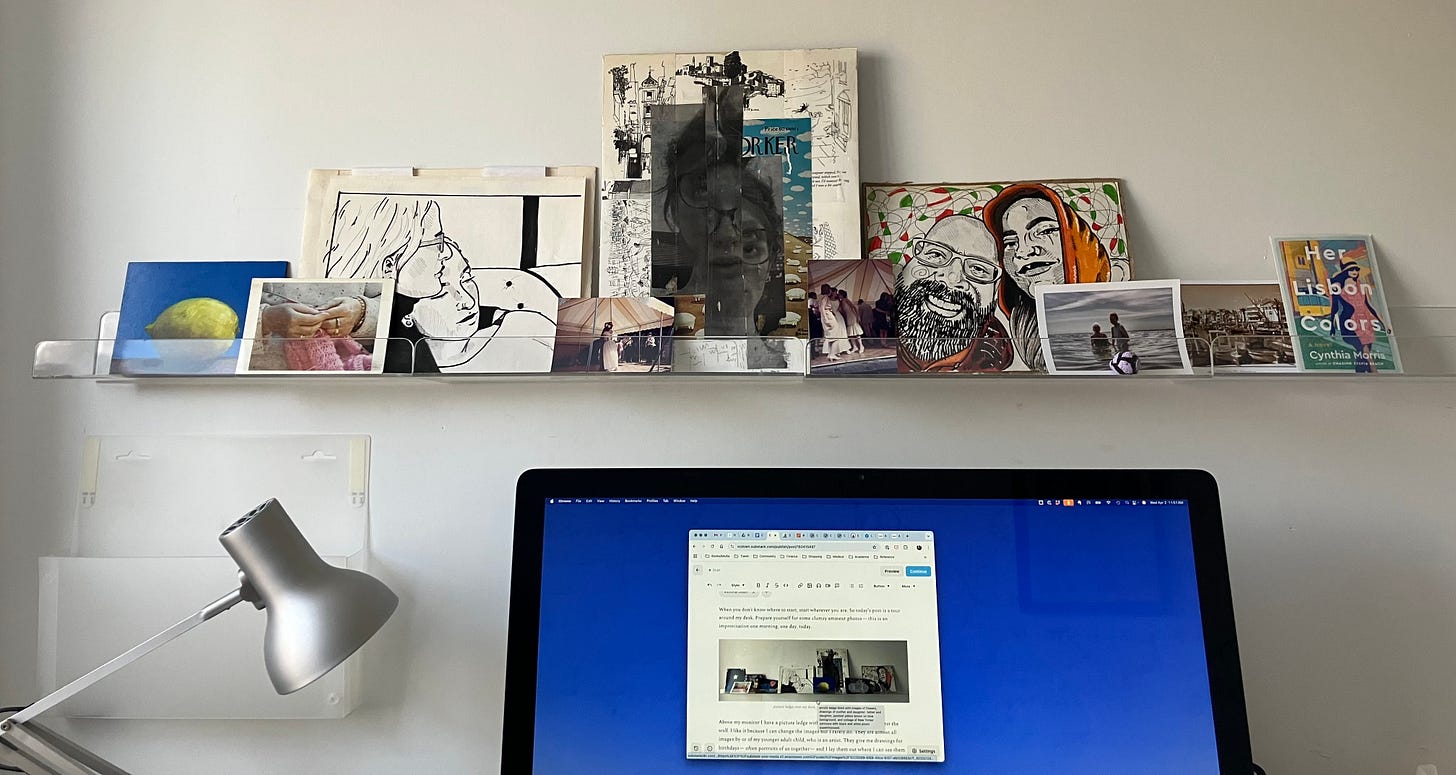 picture ledge with assorted photographs, art, and postcards over desk with blue monitor and reading lamp