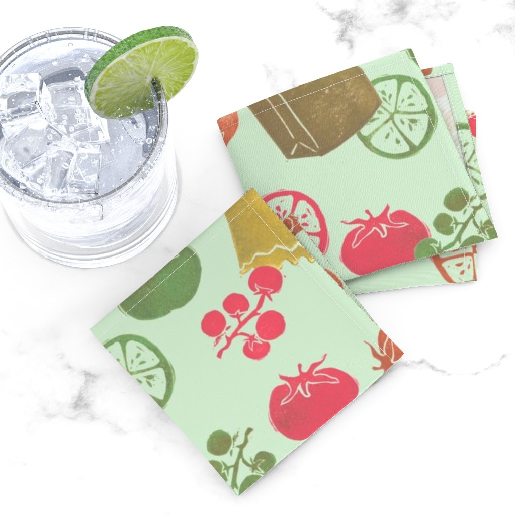 Top down of two folded Cocktail Napkins overlapping next to a glass tumbler full of ice and liquid with a lime wedge on a white marble tabletop