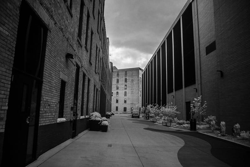 Black-and-white photograph of a downtown Kalamazoo alley between brick buildings and a modern government structure, symbolizing the space between institutions and everyday life.
