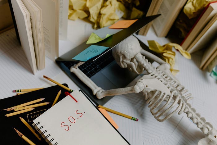 A skeleton collapsed in front of a laptop while there’s an “SOS” alert in the notebook to the side
