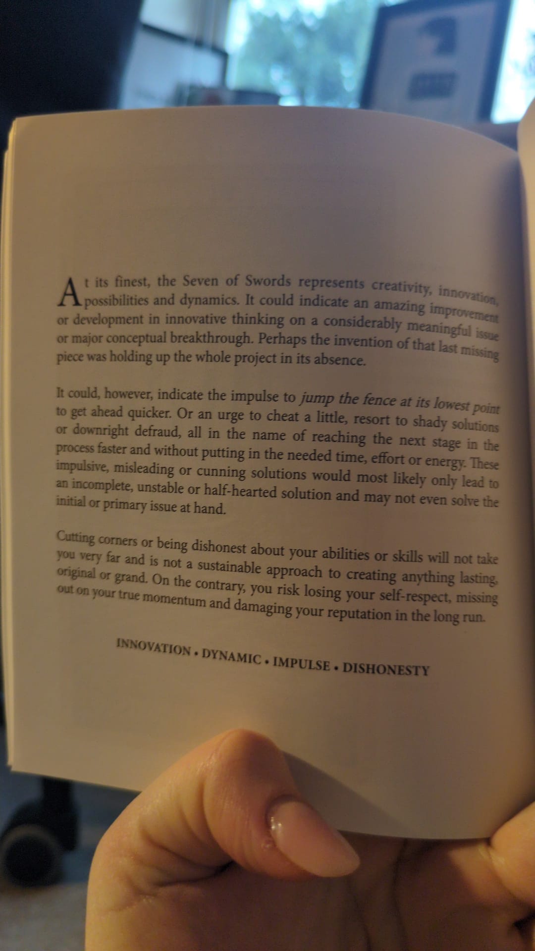 A photograph of an open tarot guidebook page interpreting the Seven of Swords. The text discusses creativity, innovation, impulse, and the risks of cutting corners or acting dishonestly, contrasting genuine progress with short-term gains. At the bottom of the page, the keywords “INNOVATION · DYNAMIC · IMPULSE · DISHONESTY” are printed.