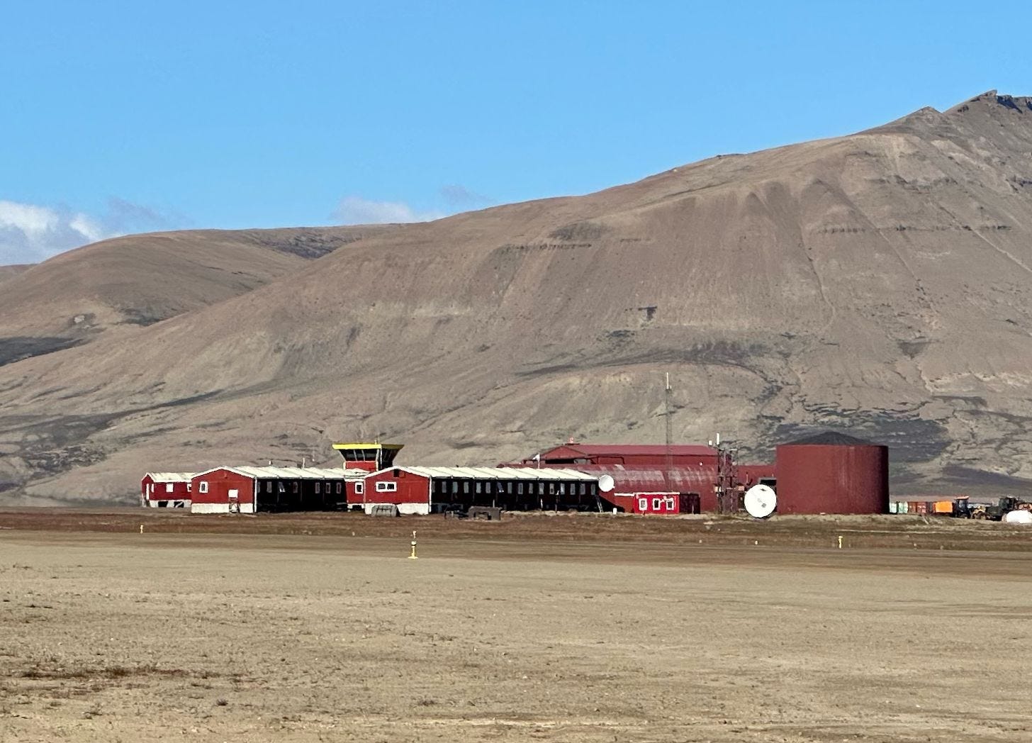 A distant view of Nerlerit Inaat airport. A long low red building with white roof and a small yellow control tower sit against a large dun-colored mountain. Flat gravel terrain in the foreground. Blue sky above.