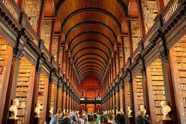 The Long Room in the Trinity College Dublin Old Library. The image showcases the grand hall with its high, vaulted wooden ceiling and rows of tall, dark wooden bookshelves filled with ancient books. Busts of notable figures line the sides of the room, and visitors can be seen walking and admiring the historic architecture and literary treasures. The atmosphere is majestic and scholarly, reflecting the library’s rich heritage.