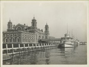 Black and white photo of the enormous immigration Station at Ellis Island. A brick building with copper topped towers and huge arches with windows. There is a ferry docked at the station. The picture was taken circa 1902 Black and white photo of the enormous immigration Station at Ellis Island. A brick building with copper topped towers and huge arches with windows. There is a ferry docked at the station. The picture was taken circa 1902