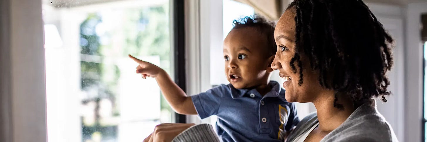 A smiling woman with dark, curly hair holds a baby boy by a bright window. The baby, wearing a navy polo shirt, points eagerly toward something outside while looking out with wide eyes and an open mouth, as if speaking. Greenery is visible through the window behind them.