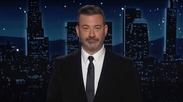 Jimmy Kimmel wearing a black suit, white shirt, and black tie, standing against a nighttime city skyline background with illuminated buildings and stars.