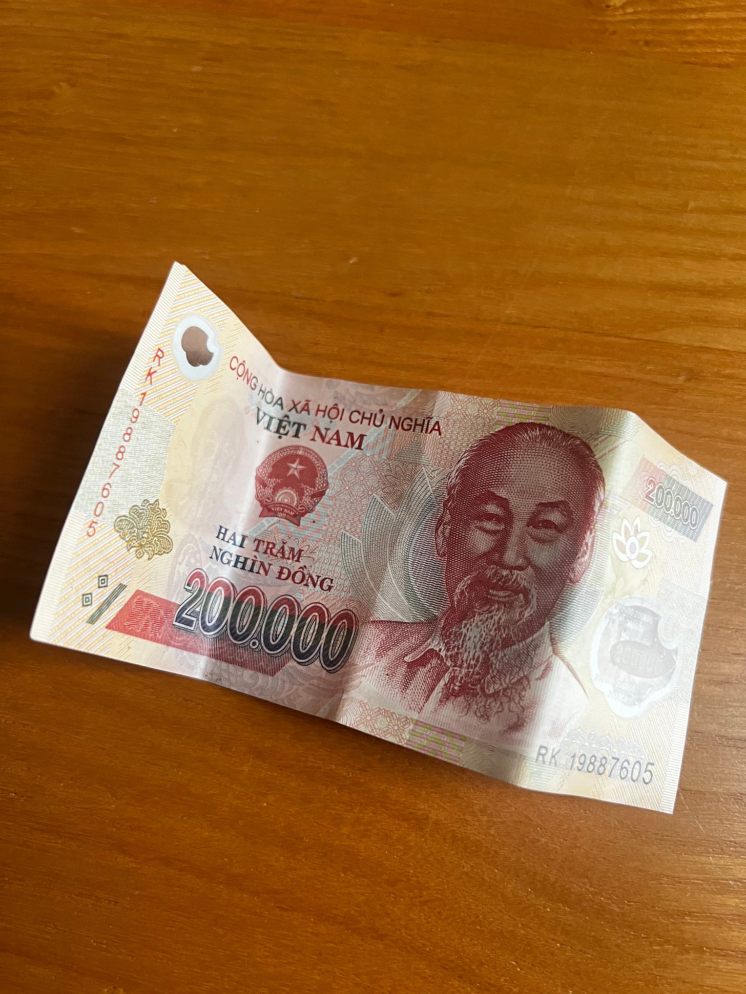 A 200,000 Vietnamese dong banknote placed on a wooden table. The polymer bill features Ho Chi Minh's portrait in red and cream tones, with "CỘNG HÒA XÃ HỘI CHỦ NGHĨA VIỆT NAM" (Socialist Republic of Vietnam) and "HAI TRĂM NGHÌN ĐỒNG" (two hundred thousand dong) printed in Vietnamese. The bill shows decorative patterns and the serial number.
