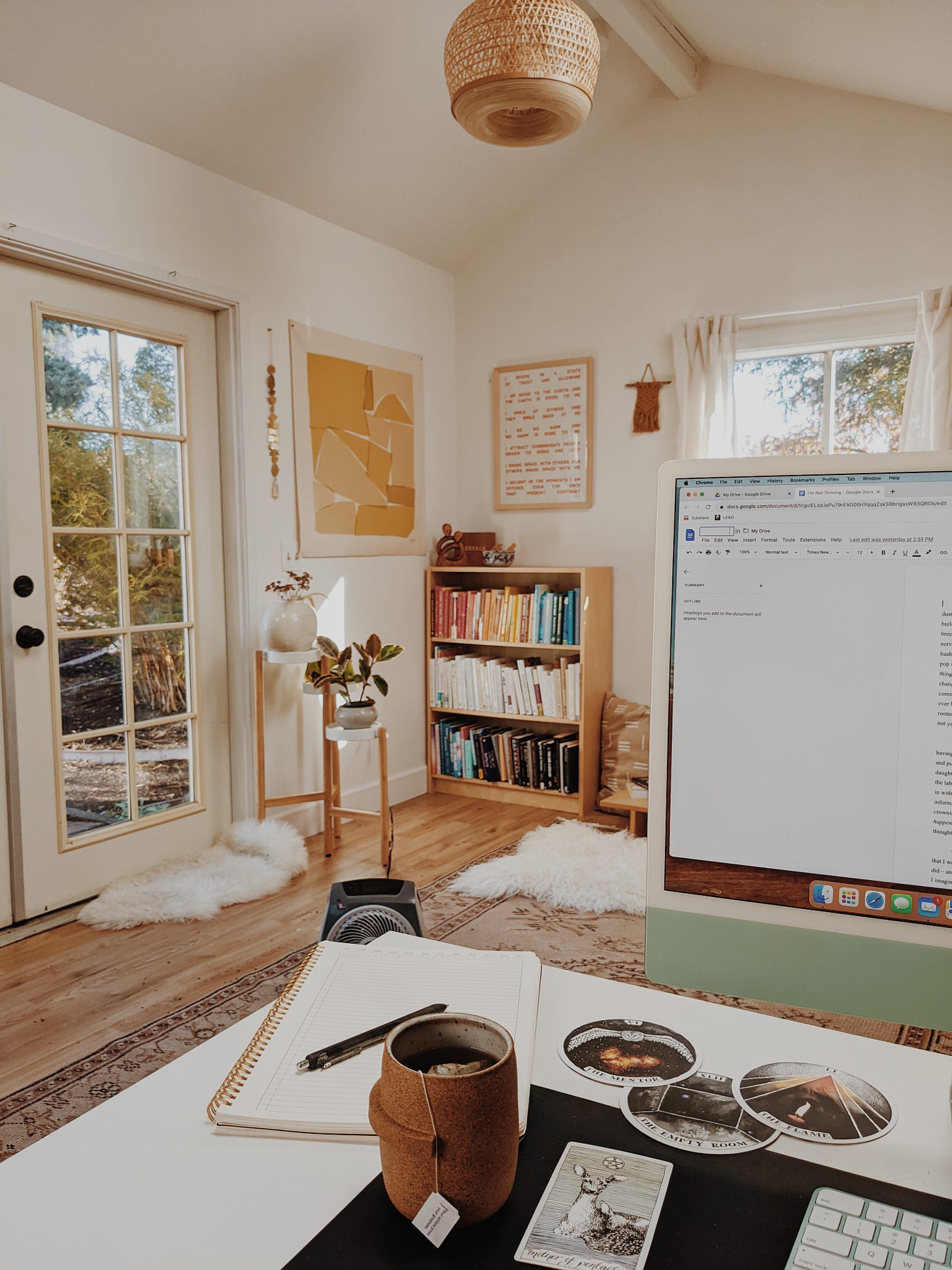 Image description: Photo of a bright, naturally-lit office space, including a cup of tea, archetype cards, a desktop computer, a bookshelf, artwork, and plants.